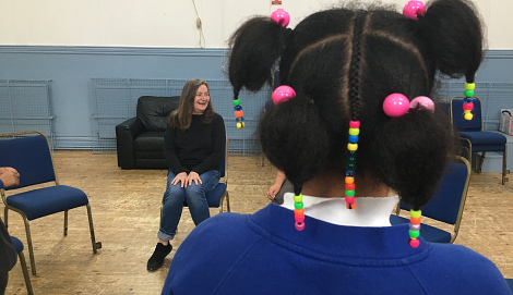 The back of a young girls head in a blue jumper with colourful beads in her hair. In the background Terry sits on a chair smiling.